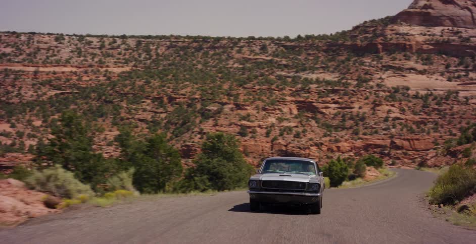 A classic silver muscle car cruises down a scenic desert road surrounded by red rock canyons