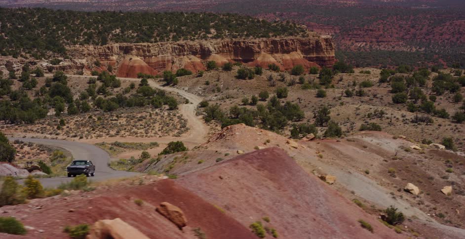 Aerial view of a vintage muscle car driving on a scenic, winding road through a vast red rock desert landscape