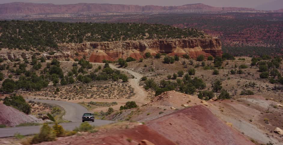 A classic Ford Mustang drives up a winding desert road surrounded by dramatic red rock canyons and vast arid landscapes