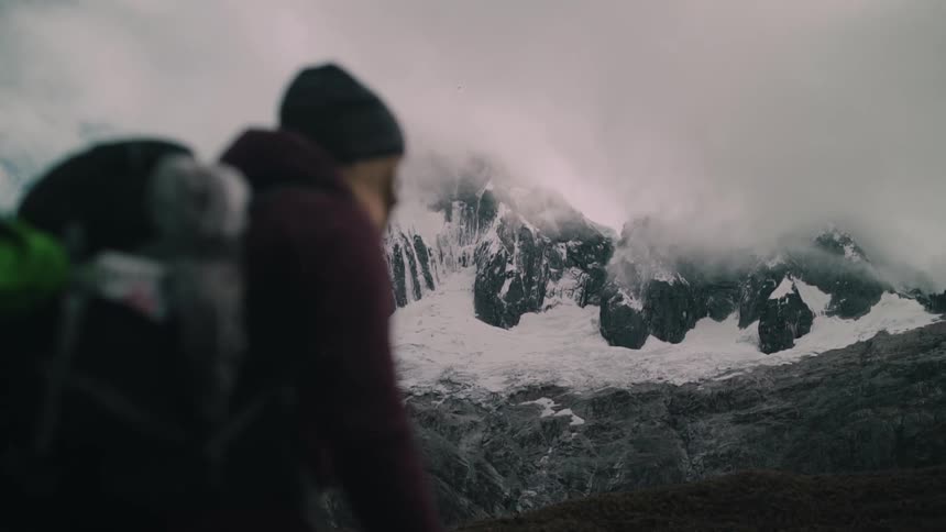 Slow-motion view of a majestic snow-capped mountain range partially obscured by clouds, with a hiker out of focus in the foreground