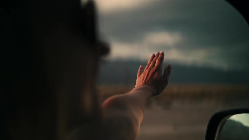 A person's hand gracefully catches the wind out of a moving car window during a cinematic sunset road trip