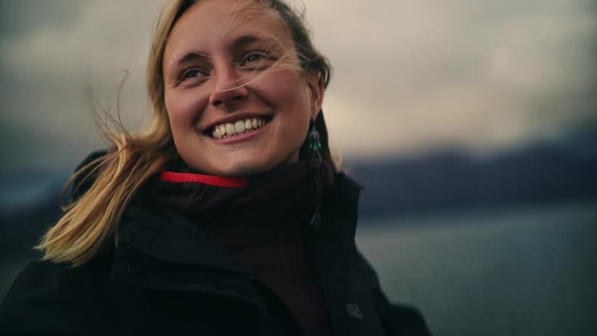 A joyful young blonde woman smiles and closes her eyes in slow motion, enjoying the feeling of the wind blowing through her hair by a mountainous lake