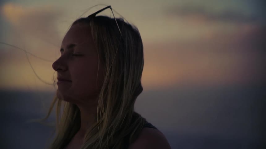 Close up profile shot of a serene young woman with eyes closed as her hair blows in the wind during a golden hour sunset