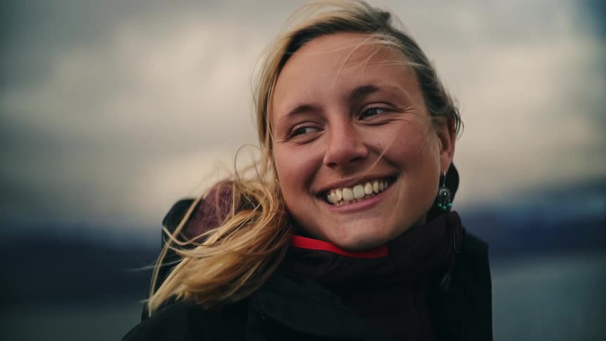 A close-up slow-motion shot of a happy young woman smiling with her eyes closed as the wind blows through her blonde hair outdoors