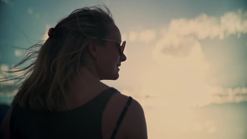 A thoughtful woman with windblown hair is silhouetted against a dreamy, bright sky with cloud reflections