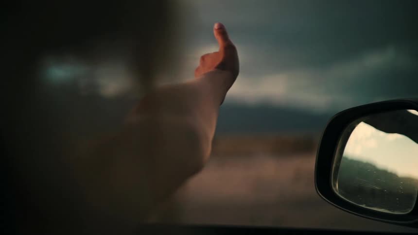 A free-spirited person's arm hangs out the window of a moving car, their hand catching the wind in slow motion during a road trip at sunset