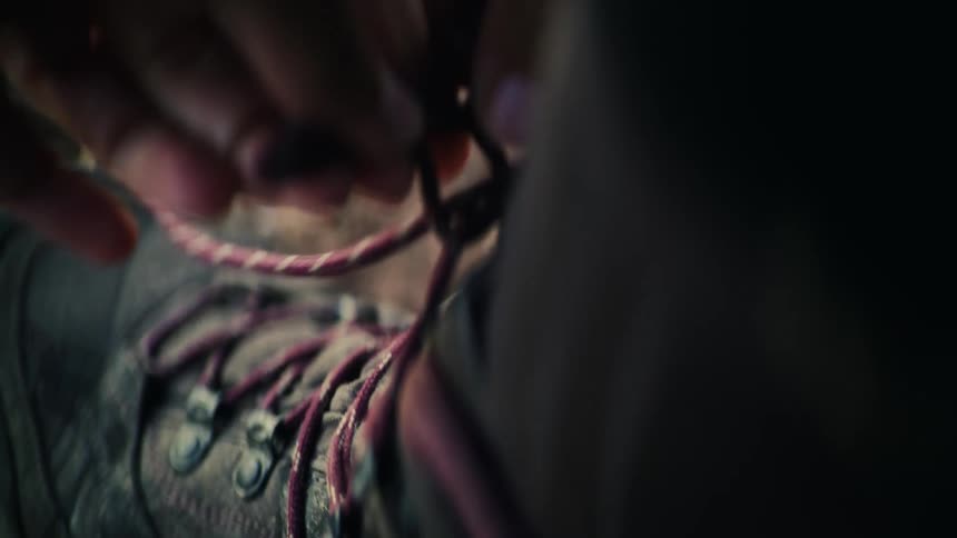 A hiker carefully ties the shoelaces of their sturdy hiking boot in a cinematic slow-motion close-up