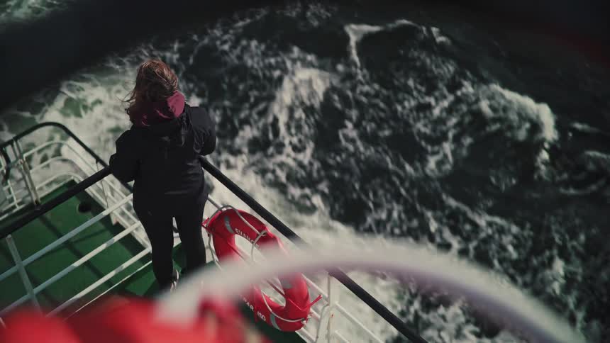 A woman stands at the railing of a moving ferry, watching the churning waves as the wind blows through her hair