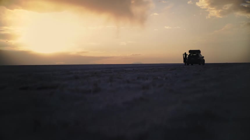 A lone traveler gets out of their overlanding vehicle to watch the golden sunset over the vast salt flats