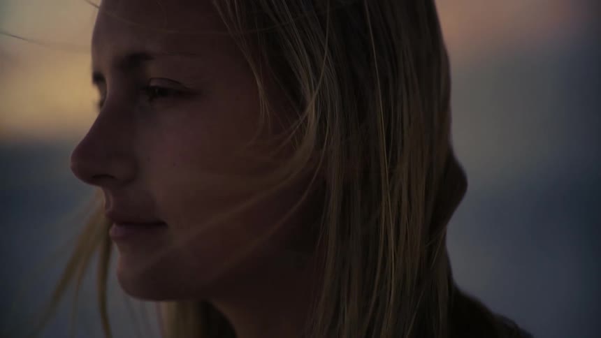 A close-up profile of a young blonde woman's face, her eyes closed in serene contemplation as her hair blows gently in the evening wind