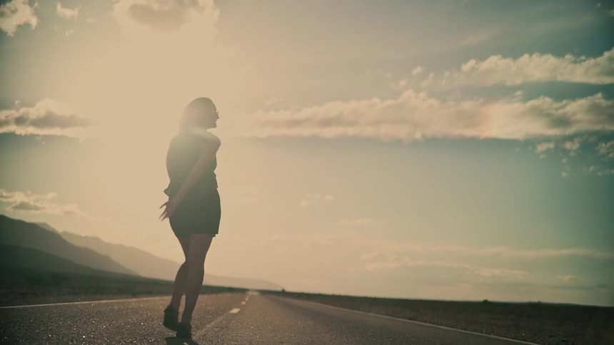 Slow motion silhouette of a woman raising her arms in freedom and walking down the middle of a desolate desert road at sunset
