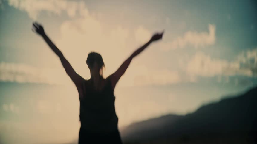 A woman raises her arms in freedom on a deserted road, silhouetted against the bright, setting sun in slow motion