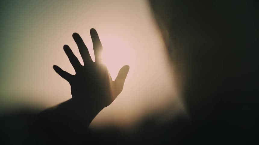 The silhouette of a hand reaching towards a bright light as rays of sun stream through the fingers