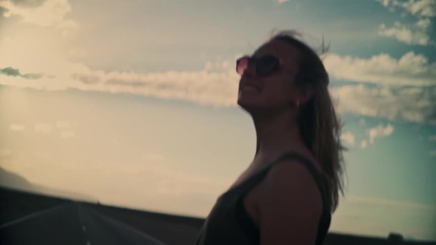 A smiling woman with sunglasses enjoys the warm sunset glow on a deserted road as the wind blows through her hair in slow motion