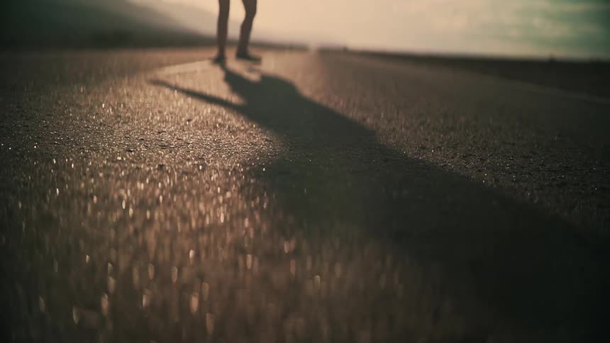 Low angle shot of a person's long shadow as they walk and dance down an empty desert road at sunset