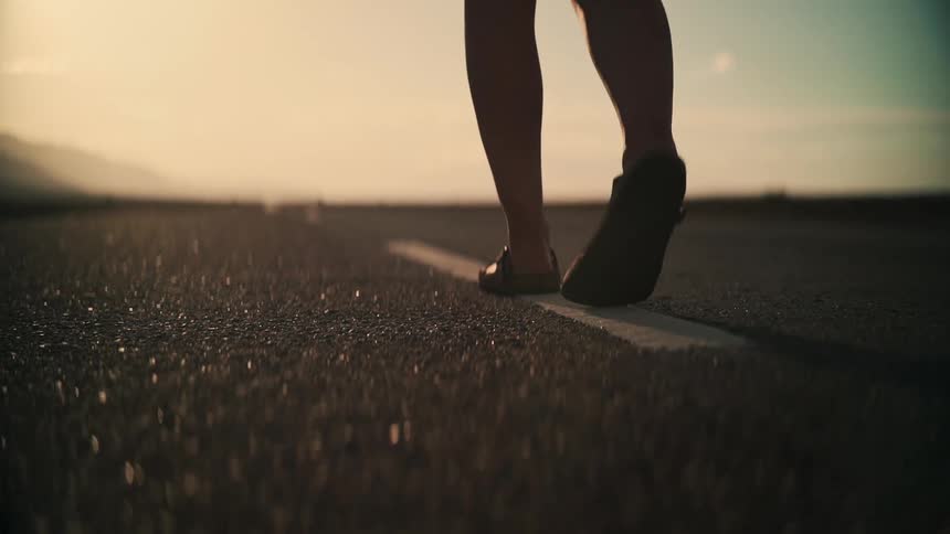 Low angle slow motion shot of a woman's feet walking down the white line of a desolate highway at sunset
