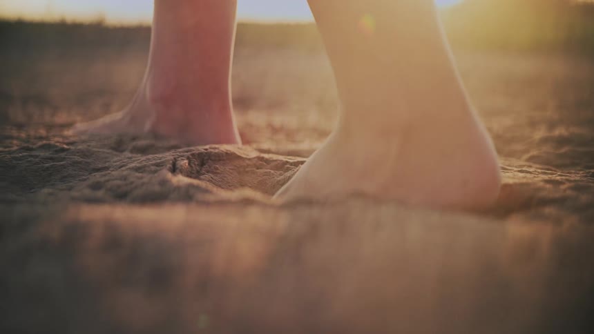 A slow-motion, low-angle closeup shows a person's bare feet walking through soft, golden sand at sunset