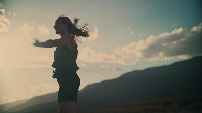A carefree young woman in silhouette twirls and dances happily with her arms outstretched in the warm glow of a mountain sunset
