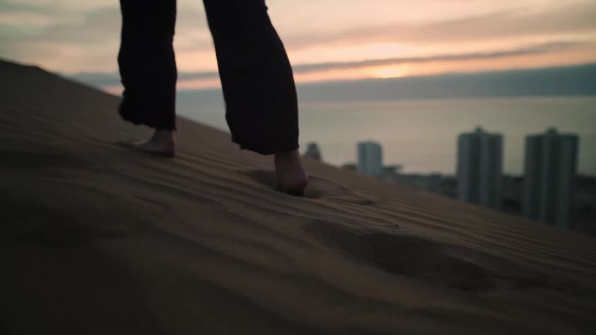 A person walks barefoot in slow motion up a sand dune carrying their shoes, with a coastal city skyline visible at sunset