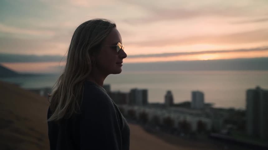 A serene woman watches the sunset over a coastal city with her hair blowing in the wind in slow motion