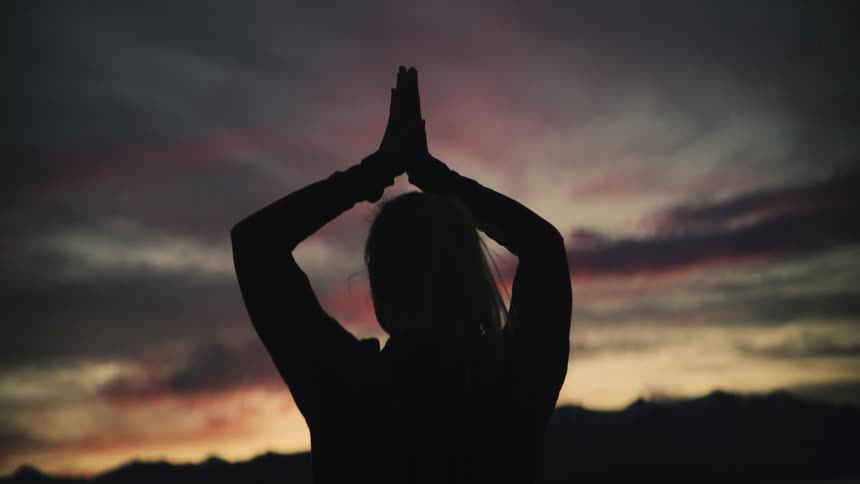 A woman's silhouette slowly raises her arms into a prayer pose against a dramatic and colorful sunset sky