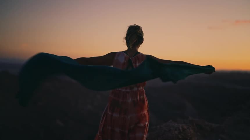 A free-spirited woman seen from behind holds a long, flowing scarf that catches the wind against a stunning sunset sky in slow motion