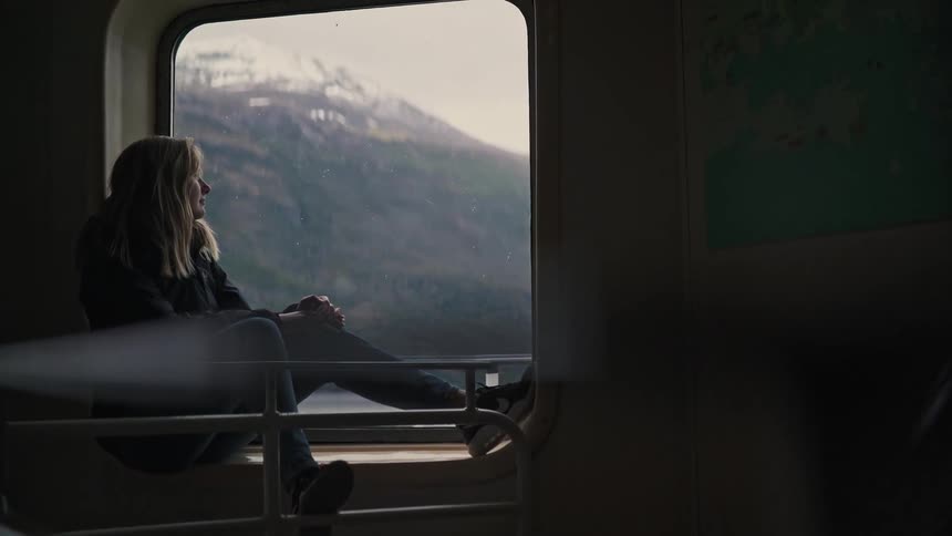 A woman sits contemplatively by the window of a ferry, watching the majestic snow-capped mountains drift by