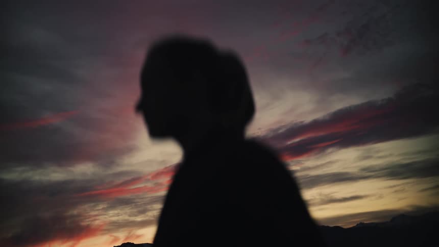 The silhouette of a contemplative woman slowly stretches her arms towards the dramatic sunset sky over a mountain lake