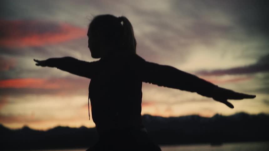 A woman's silhouette holds a Warrior II yoga pose in slow motion against a dramatic sunset sky over a mountain lake