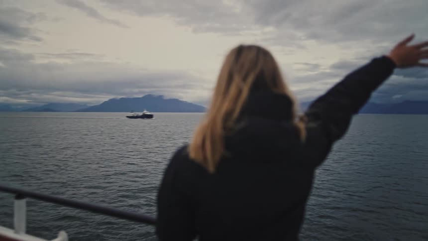 A woman on a ferry waves goodbye to a passing ship on a vast, moody sea with mountains on the horizon