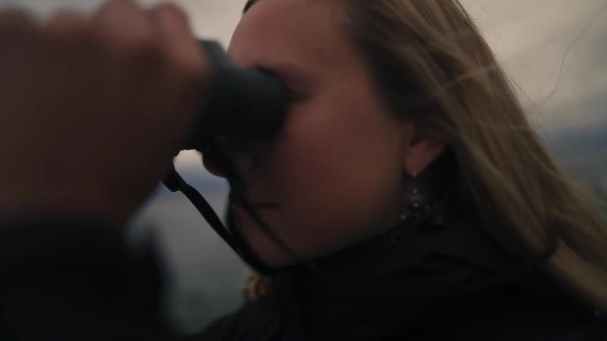 A slow-motion close-up of a young woman looking through binoculars while her hair blows in the wind