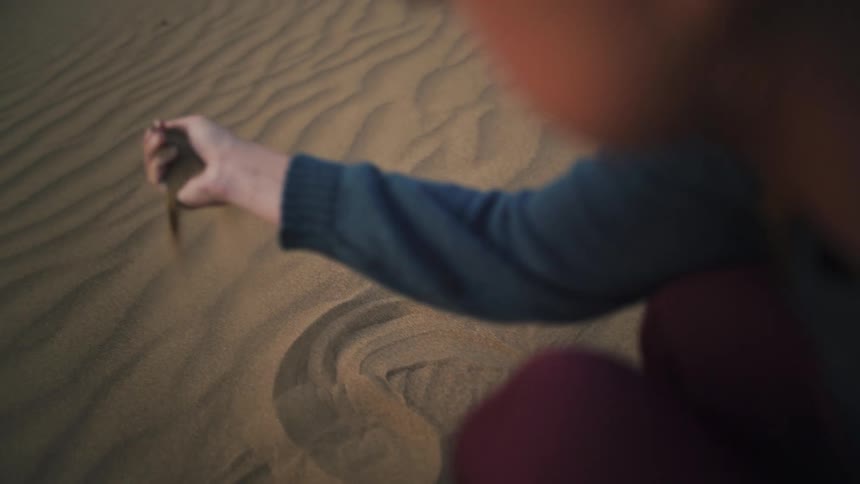 An over-the-shoulder slow-motion shot of a woman letting soft desert sand fall through her fingers