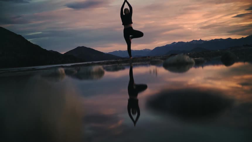 The silhouette of a woman in a tree yoga pose is perfectly reflected in a still mountain lake during a beautiful sunset