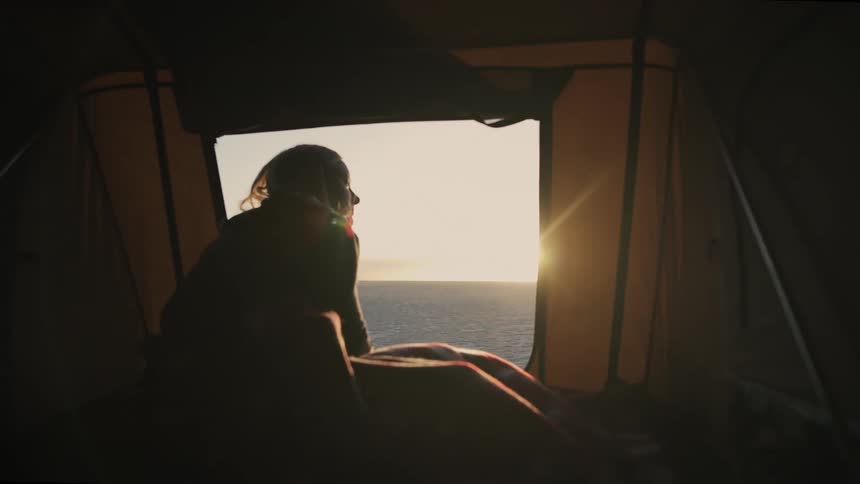 A silhouetted woman sits in a cozy tent and watches the golden sun rise over the calm ocean
