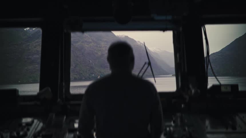 A silhouetted captain stands on the bridge of a ship, looking out the window at the majestic mountains of a fjord