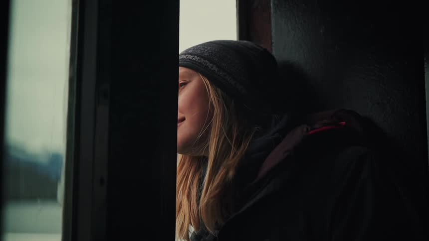A young woman in a beanie smiles as she looks out the window of a boat at the passing scenery in slow motion