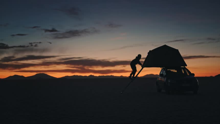 Silhouette of a person climbing a ladder into a rooftop car tent in the vast desert at sunset