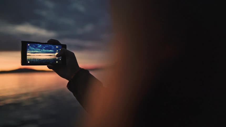 Over-the-shoulder shot of a person's silhouette as they capture a beautiful sunset over the calm ocean on their smartphone