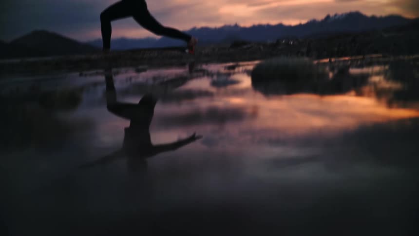 A woman's silhouette is reflected in the water as she holds a warrior yoga pose on a lakeshore during a beautiful sunset with mountains in the background