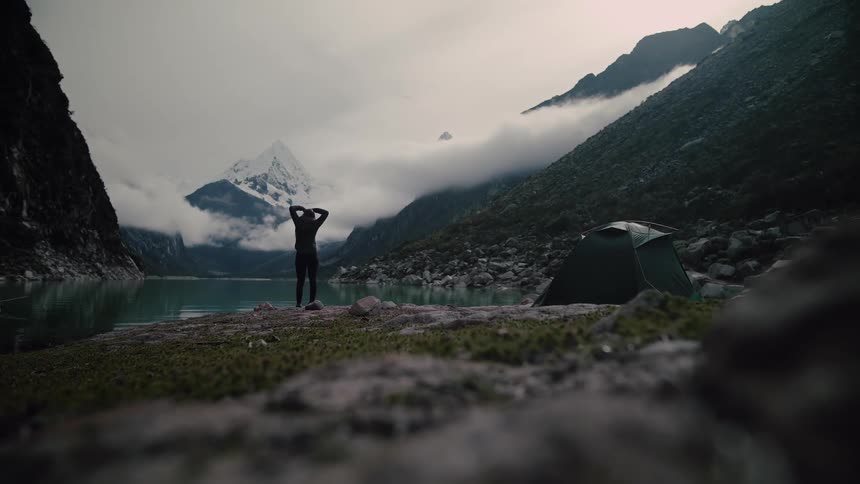 A female adventurer exits her tent, walks to the edge of a pristine mountain lake, and stretches with her arms raised, surrounded by epic, snow-capped peaks