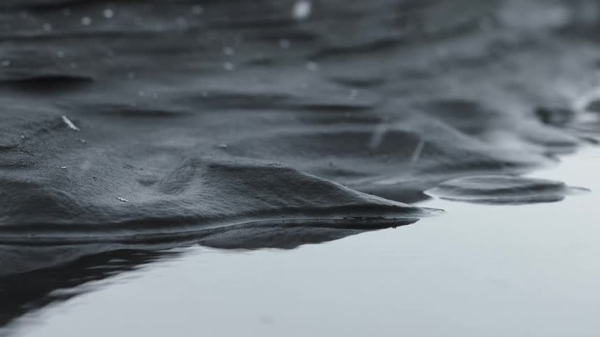 A serene, black and white close-up shows light snow falling gently onto the calm surface of the water