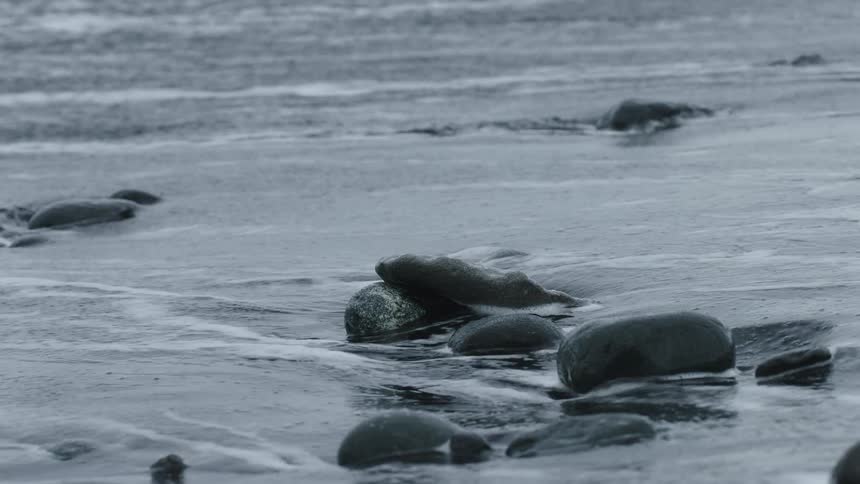 Slow motion shot of foamy ocean water receding over dark stones on a black sand beach
