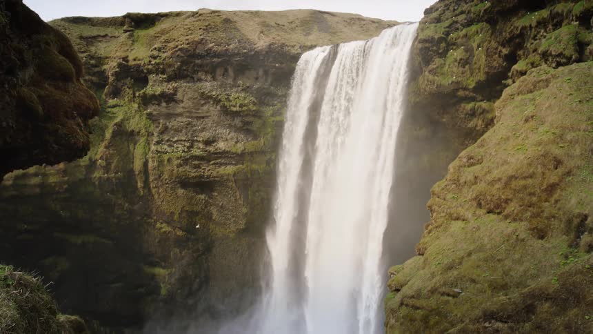 A slow tilt down reveals the majestic Skogafoss waterfall in Iceland as it plunges over mossy cliffs into a misty pool below