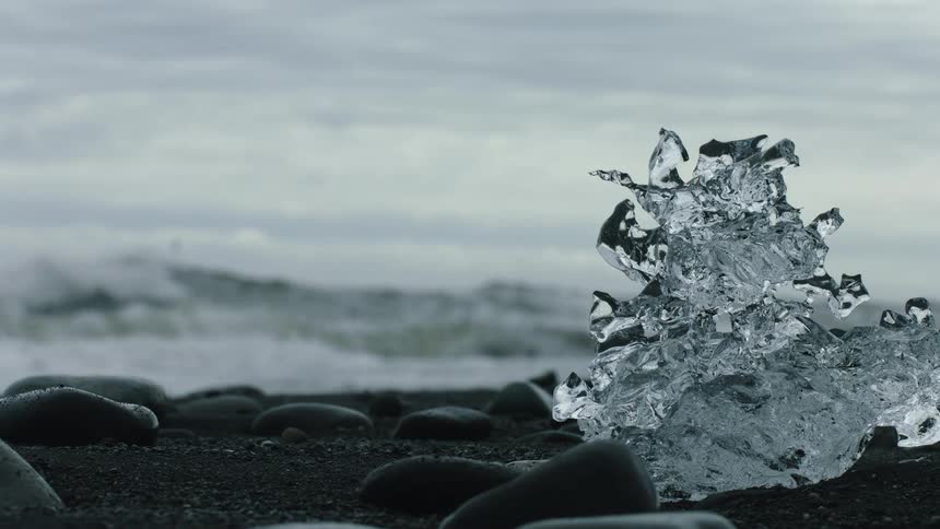 A sculpted shard of glacial ice rests on a black sand beach as ocean waves gently roll ashore in the background