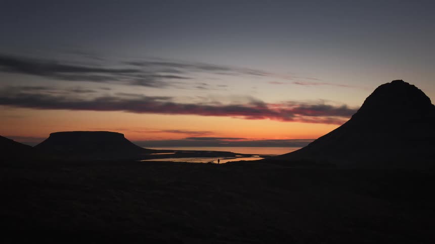 A lone traveler stands amidst a vast Icelandic landscape, silhouetted against the vibrant colors of a sunset over the water and mountains