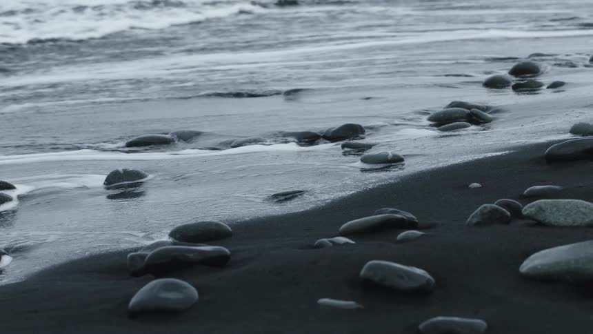 Gentle ocean waves in slow motion wash over smooth black stones on a dark volcanic sand beach