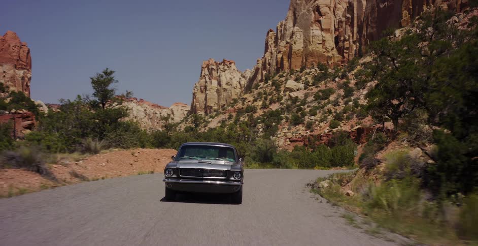 A classic silver muscle car cruises down a winding desert road surrounded by towering red rock formations