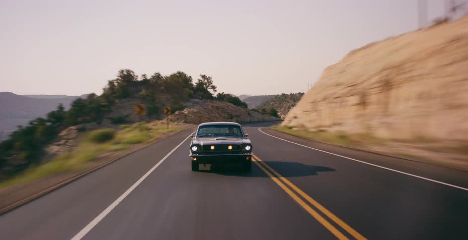A classic black muscle car speeds along a scenic and winding desert highway at sunrise