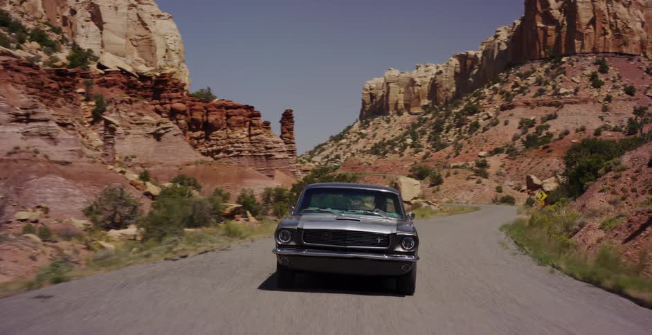 A classic grey Ford Mustang cruises down a winding desert road surrounded by dramatic red rock canyon walls on a sunny day