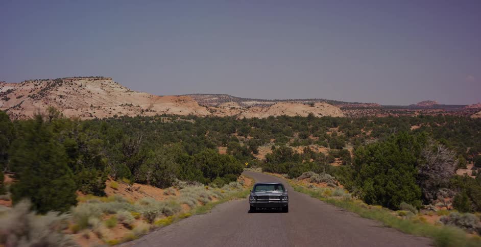A classic black muscle car drives on a remote desert highway through a scenic and rugged landscape